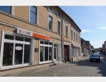 Straßenansicht mit einer Reihe von historischen, hellbraunen Gebäuden mit weißen Fensterrahmen und dekorativen Fassadenelementen; im Vordergrund ein Friseursalon mit einem orangefarbenen Schild "Haar Studio" und großen Schaufenstern. Die gepflasterte Straße führt an geparkten Autos entlang durch ein ruhig wirkendes Stadtviertel bei sonnigem Wetter.