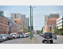 Straßenszene in einer Stadt mit zwei Fahrspuren in jede Richtung, gesäumt von mehrstöckigen Wohn- und Bürogebäuden. Im Vordergrund sind Autos in einem Stau zu sehen, Laternenmasten trennen die Fahrbahnen; im Hintergrund ragen moderne Wohnhochhäuser auf.