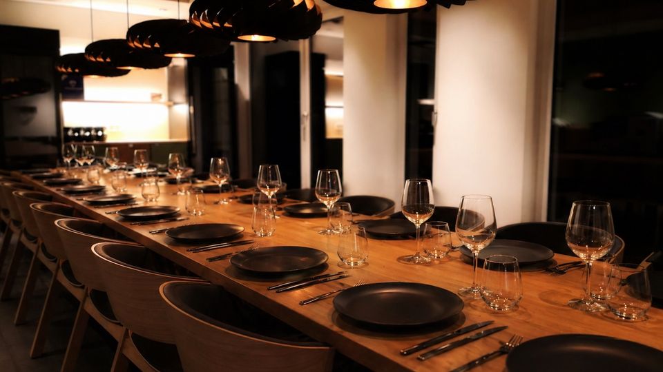 Moderner Speisebereich mit einem langen Holztisch, gedeckt mit schwarzen Tellern, Besteck und mehreren Wein- und Wassergläsern. Über dem Tisch hängen große schwarze Pendelleuchten, dazu Holzdecke und große Fenster in einem offenen Raum mit angrenzender Küche.