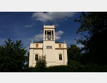 Ein helles, historisches Gebäude mit rechteckigem Turm und Glocken im oberen Bereich, umgeben von Bäumen und Sträuchern. Der Himmel ist klar mit ein paar Wolken im Hintergrund.