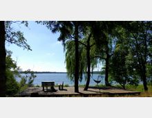 Holzplattform am Ufer eines Sees, umgeben von Bäumen, darunter eine hängende Weide. Auf der Plattform befinden sich eine Sitzgruppe mit einem Tisch und eine Hängematte zwischen zwei Bäumen, der See erstreckt sich im Hintergrund.