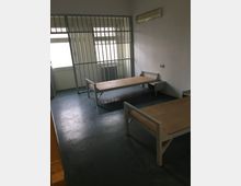 Spartanischer Innenraum mit zwei einfachen Metallbetten und kleinem Nachttisch auf dunklem Boden. Am Fenster befindet sich ein Gitter mit Tür, dahinter Heizkörper und Tageslicht; an der Wand ist eine Leuchtstofflampe montiert.
