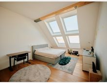 Schlafzimmer in einer Dachgeschosswohnung mit schräger Wand und großen Dachfenstern, die viel Tageslicht hereinlassen. Der Raum enthält ein graues Bett, einen Schreibtisch mit Stuhl, einen Teppich auf Holzboden sowie eine Kommode mit dekorativen Gegenständen.