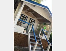 Kleines weißes Gartenhaus mit blauen Fensterrahmen und einem Vordach aus Holz, das mit einem Schilfgeflecht verkleidet ist. Eine Holztreppe mit blau-weißen Geländern führt zur überdachten Veranda, die mit Korbmöbeln und Pflanzen dekoriert ist.