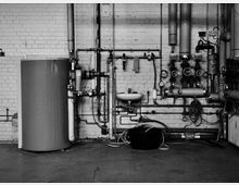 Schwarz-weiß-Foto eines technischen Raums mit einer Backsteinwand. Zu sehen sind ein großer Boiler, Rohre, Ventile, Druckanzeigen und ein Waschbecken sowie Verlängerungsschläuche und ein schwarzer Eimer auf dem Boden.