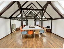 Großer Besprechungsraum in einem Dachgeschoss mit sichtbaren dunklen Holzbalken, hellen Wänden und Holzfußboden. Ein rechteckiger Tisch ist mit bunten Stühlen umgeben, und im Hintergrund befinden sich eine Flipchart, ein Monitor und niedrige Holzmöbel.