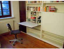 Arbeitszimmer mit großem weißen Einbauregal voller Bücher und einer ausklappbaren Schreibtischplatte. Daneben ein Fenster mit Heizkörper darunter sowie ein Holzfußboden und ein Bürostuhl vor dem Regal.