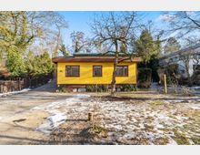 Einstöckiges, gelb gestrichenes Wohnhaus mit flachem Dach und breiten Fenstern, umgeben von Bäumen und Sträuchern. Davor liegt ein offener Hof mit Zufahrt und stellenweise schneebedecktem Boden.