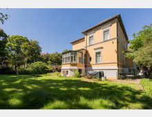 Zweistöckige Villa mit gelber Fassade und weißen Fensterrahmen, mit einem verglasten Erkeranbau an der Seite. Davor liegt ein großer, grasbewachsener Garten mit Bäumen und Sträuchern, neben einem kleinen eingezäunten Bereich am Haus.