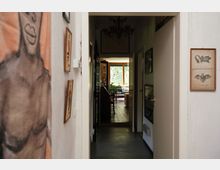 Ein schmaler Flur in einer Wohnung, geschmückt mit Wandbildern und kleinen Rahmen an den Wänden. Der Flur führt in ein Wohnzimmer mit großen Fenstern, die Tageslicht hereinlassen, Holzboden und Pflanzen im Hintergrund.