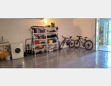 Ordentliche Garage mit grauem, glänzendem Boden. An der linken Wand befinden sich eine Waschmaschine, ein Waschbecken und Regale mit Werkzeugen und Haushaltsgegenständen; rechts stehen drei Fahrräder, und eine geöffnete Tür führt ins Freie.