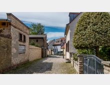 Schmale, gepflasterte Gasse in einem Dorf mit historischen Gebäuden aus Backstein und Putzfassaden. Rechts ein gepflegter Garten mit einem großen, geschnittenen Baum und einem Metallzaun, links alte, verwitterte Backsteinmauern, im Hintergrund farbige Häuser mit roten Ziegeldächern.