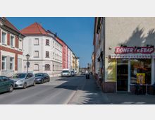 Straßenszene in einem Wohnviertel mit mehrstöckigen, farbigen Gebäuden im historischen Stil auf der linken Seite und einem Döner-Imbiss mit rotem Schild und Markise auf der rechten Seite. Geparkte Autos und Transportfahrzeuge säumen die gepflasterte Straße, während der Himmel klar und blau ist.