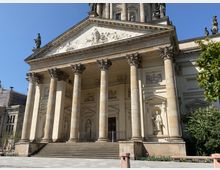 Historisches Gebäude mit klassizistischer Architektur, geprägt durch eine Monumentalfront mit sechs korinthischen Säulen, aufwendig verzierten Giebelreliefs und Statuen in Nischen. Im Vordergrund führen breite Steinstufen zu einem überdachten Eingang, seitlich befinden sich Bäume und ein gepflasterter Platz.