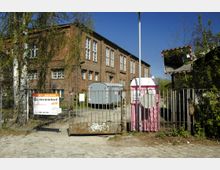 Backsteingebäude mit großen Fenstern, hinter einem Metallzaun mit einem „Zu Vermieten!“-Schild. Im Vordergrund ein rosa mobiles Toilettenhäuschen und ein Verkehrsspiegel; der Boden ist gepflastert, umgeben von Bäumen und Sträuchern.