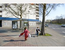Straßenszene mit einem Gehweg, auf dem zwei Kinder laufen, sowie einer Bushaltestelle mit einem Werbeposter. Im Hintergrund befinden sich ein modernes, weißes Gebäude mit blauer Fassade sowie eine mehrspurige Straße mit parkenden und fahrenden Autos.