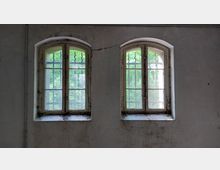 Zwei alte, leicht verschmutzte Fenster mit Rundbögen in einer weißen, abgeblätterten Wand; die Fenster haben Gitterstäbe und ermöglichen den Blick auf grüne Vegetation draußen.