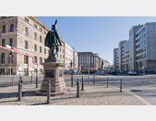 Eine Stadtansicht mit einer bronzenen Statue auf einem Podest im Vordergrund, umgeben von gepflastertem Boden und Pollern mit Ketten. Im Hintergrund stehen historische und moderne Gebäude entlang einer breiten Straße unter klarem Himmel.