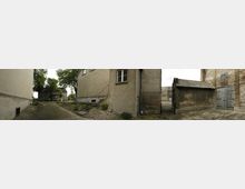 Panorama einer ländlichen Hofsituation mit alten Gebäuden aus verputztem Stein und Ziegeln. Links und in der Mitte sind Wohnhäuser mit einfachen Fenstern und Außentreppen zu sehen, rechts ein Nebengebäude mit schlichten Holztüren; der Hof ist teils begrünt, teils betoniert.