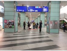 Eingangsbereich eines Bahnhofs mit blauem Schild, das den Ausgang „Friedrichstraße“ und weitere Hinweise zu U-Bahn, WC und Taxis anzeigt. Der Bereich ist von hellgrünen Glaswänden umgeben, mit Werbeplakaten, Automaten und verschiedenen Personen, darunter Passanten und ein Hund.