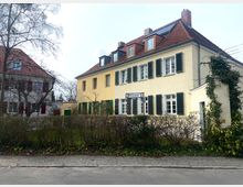 Zwei benachbarte Wohnhäuser im traditionellen Stil, eines mit gelber Fassade, grünen Fensterläden und rotem Ziegeldach, das andere mit grauer Fassade, roten Fensterläden und ähnlichem Ziegeldach. Im Vordergrund wächst eine Hecke, und ein Straßenschild mit der Aufschrift "Rumeyplan" ist sichtbar.