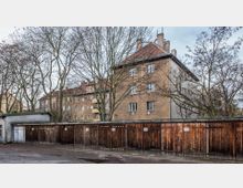 Mehrstöckiges Wohnhaus mit einfacher Fassade aus bräunlichem Putz, im Vordergrund eine Reihe von hölzernen Garagen mit vereinzelt angebrachten Schildern. Karge, unbelaubte Bäume stehen zwischen den Gebäuden, und der Boden ist teilweise nass.