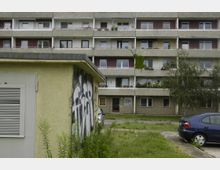 Wohnanlage mit einem mehrstöckigen Gebäude aus Beton, Balkonen und begrünten Fassadenelementen im Hintergrund. Im Vordergrund steht ein kleines, gelb gestrichenes Gebäude mit Graffiti, daneben befindet sich eine gepflasterte Fläche mit parkenden Autos und angrenzendem Gras.