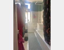 Ein kleines Badezimmer mit weißen Fliesen und grün gestrichenen oberen Wänden. Links hängen Handtücher, rechts befindet sich eine Badewanne mit einem Vorhang mit Blumenmuster. Vorne stehen ein Waschbecken und eine Toilette, darüber ein Fenster und ein eingerahmtes Poster.