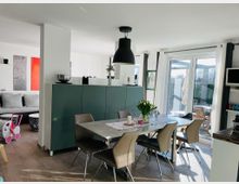 Moderne Wohnküche mit offenem Essbereich, bestehend aus einem rechteckigen Esstisch mit sechs Stühlen und einer Hängelampe darüber. Im Hintergrund sind eine graue Couch, eine halbhohe grüne Trennwand und große Fenstertüren sichtbar, die zu einer Terrasse mit Gartenblick führen.