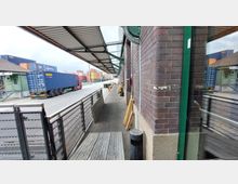 Industriegebiet mit Lagerhallen: Ein Gehweg neben einer Backsteinfassade mit Vordach, das von Stahlträgern gestützt wird. Im Hintergrund sind Container, ein geparkter LKW und der Zaun eines Containerdepots sichtbar.