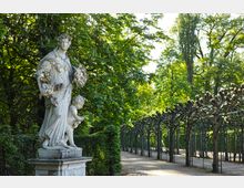 Marmorstatue einer Frau mit Obstkorb und einem Kind auf einem Sockel, umgeben von grüner Vegetation; daneben verläuft ein überdachter Laubengang aus beschnittenen Bäumen in einem Park.