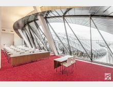 Innenansicht eines modernen Konferenzraums mit einer Glasfassade, die eine geschwungene, kuppelartige Außenstruktur zeigt. Der Raum hat rote Teppichböden, weiße Stühle und rechteckige Tische, mit einem Blick auf eine futuristische Architektur im Hintergrund.