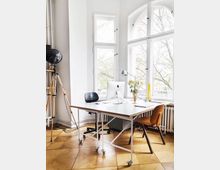 Bright office workspace with a large white desk on casters, two chairs, and a desktop computer, set on a parquet wood floor. Tall arched and rectangular windows fill the wall, with a vintage tripod floor lamp and a radiator beneath the windows.