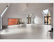 Minimalistischer, heller Innenraum mit weißem Boden und spitz zulaufender Decke, geprägt von großen, asymmetrischen Fenstern, die rote Backsteinmauer und Stadtansichten zeigen. Der Raum ist spärlich möbliert mit wenigen Stühlen, einem Tisch und einer Vase mit Blumen.