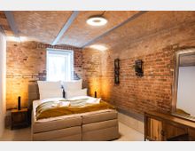 Schlafzimmer mit freiliegenden Ziegelwänden und einem gewölbten Ziegeldecken, ausgestattet mit einem neutralen Polsterbett, Nachttischen aus Holz, einem großen Spiegel und minimalistischen Pflanzenregalen. Ein Fenster sorgt für Tageslicht, während eine moderne Deckenleuchte den Raum zusätzlich beleuchtet.