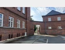 Backsteinmauergebäude mit Fenstern und verzierten Ziegelsteindetails, verbunden durch einen Torbogen mit einem Gittertor. Im Vordergrund befinden sich Parkplätze, im Hintergrund sind Bäume sichtbar.