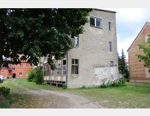 Ein altes, verputztes Gebäude mit teils beschädigten Wänden und verglasten Holzkonstruktionen, umgeben von Grasflächen, Bäumen und weiteren Gebäuden aus rotem Ziegel und Putz. Im Hintergrund sind moderne Wohnhäuser und parkende Autos zu sehen.