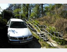 Ein geparktes weißes Auto steht neben einer schmalen Außentreppe mit Metallgeländern, die einen zugewachsenen Hang hinaufführt. Im Hintergrund sind Bäume, Sträucher und ein Maschendrahtzaun zu sehen.