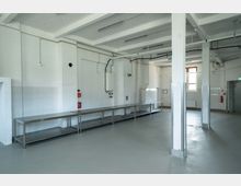 Großer, heller Raum mit weißen Wänden und Decke aus Beton mit sichtbaren Trägern. Der Boden ist grau und glatt, entlang der Wände stehen lange, metallene Arbeitstische; an den Wänden befinden sich Rohre, Steckdosen und ein Boiler.