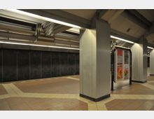 Untergrundbahnhof mit gefliesten Böden und Metallwänden, unterstützt von breiten Säulen. Eine beleuchtete Werbetafel mit Plakaten steht zwischen den Säulen, und die Decke zeigt sichtbare Metallträger.