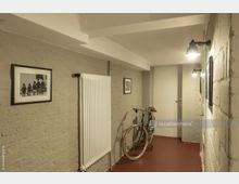 Flur mit hell gestrichenen Wänden aus Mauerwerk, an denen gerahmte Bilder hängen. Auf der linken Seite ist ein weißer Heizkörper, daneben ein abgestelltes Fahrrad; der Boden ist rot, und die Wände werden von Wandlampen beleuchtet.