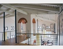 Innenansicht eines modernen Museums oder Ausstellungsraums in einem historischen Gebäude mit offenem Dachstuhl, weißen Wänden und Rundbogenfenstern mit roten Backsteinrahmungen. Metallgeländer und Holzböden grenzen verschiedene Ebenen ab, auf denen Informationstafeln und Sitzbereiche verteilt sind.