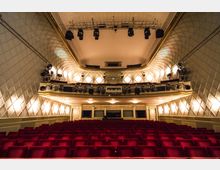 Innenansicht eines Theatersaals mit roten Polstersesseln, verzierten Wänden mit gepolsterten Paneelen und einer beleuchteten Empore. Über der Bühne befindet sich eine Lichtanlage, und mehrere Kronleuchter beleuchten den Raum.