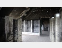 Verlassener Raum mit stark beschädigten Fliesenwänden und offener Decke mit abblätternder Farbe. Der Raum zeigt mehrere geflieste Nischen und Durchgänge, die auf eine frühere Nutzung als Nassbereich hindeuten, umgeben von sichtbarer Verfallsspuren.