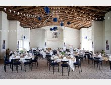 Ein rustikaler Veranstaltungsraum mit sichtbaren Holzdeckenbalken und weiß verputzten Wänden, dekoriert mit Wimpelketten und blauen Papierornamenten. Runde Tische mit weißen Tischdecken und schwarzem Stuhlset sind festlich gedeckt, umgeben von hellen Backsteinböden.