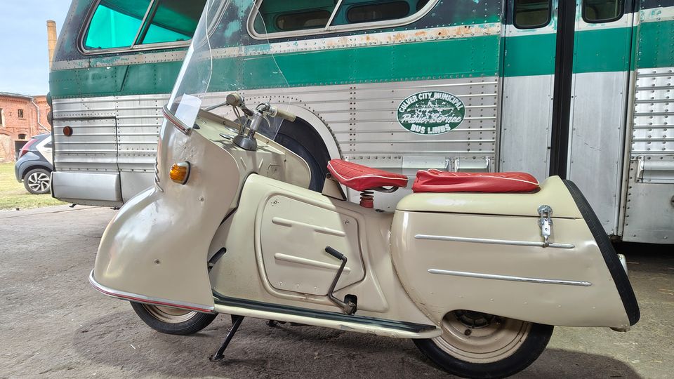 Ein cremefarbener Motorroller mit hoher Windschutzscheibe und rotem Sitz steht auf einem Betonboden unter einem überdachten Bereich. Im Hintergrund ist die Seite eines grün-silbernen, genieteten Oldtimerbusses mit großen Fenstern zu sehen, daneben ein geparktes Auto und Backsteingebäude.