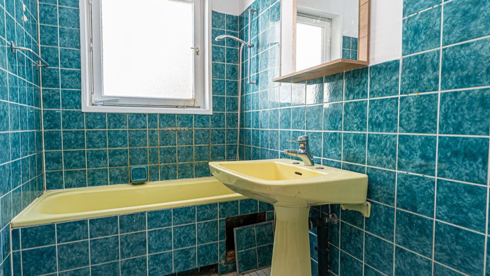 Kleines Badezimmer mit türkisblauen Wandfliesen, Badewanne mit Duscharmatur und einem gelblichen Standwaschbecken. Ein großes Milchglasfenster sowie ein Spiegel mit Ablage befinden sich über dem Waschbecken.