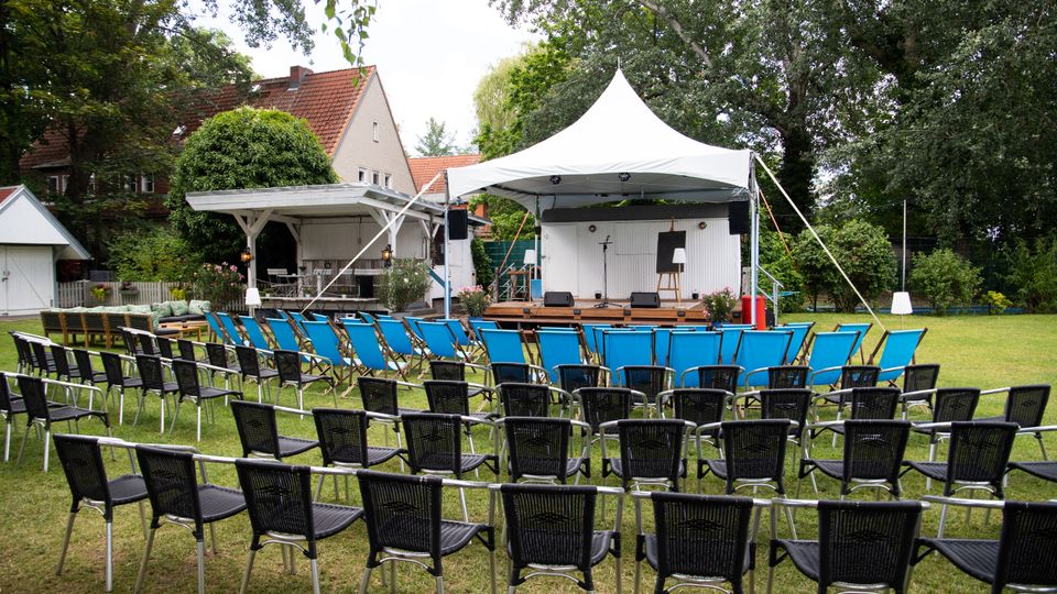 Bestuhlter Gartenbereich mit Reihen aus schwarzen und blauen Stühlen vor einer kleinen Holzbühne unter einem weißen Pavillonzelt, ausgestattet mit Mikrofonständern und Lautsprechern. Im Hintergrund stehen ein weißes Gartenhaus, ein Wohnhaus mit rotem Ziegeldach und hohe Bäume, die den Veranstaltungsplatz umgeben.