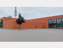 Flaches Gebäude aus roten Ziegeln mit großen Glasfenstern auf der rechten Seite und einer einzelnen Tür auf der linken Seite. Vor dem Gebäude befindet sich eine gepflasterte Fläche und ein einzelner Baum, im Hintergrund ist ein hoher Schornstein sichtbar.