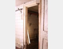 Blick in einen verlassenen, heruntergekommenen Raum mit weißen, teilweise beschädigten Wandfliesen. Die Umgebung wirkt schmutzig, und an der Wand ist eine lose, leuchtende Leuchte sichtbar.